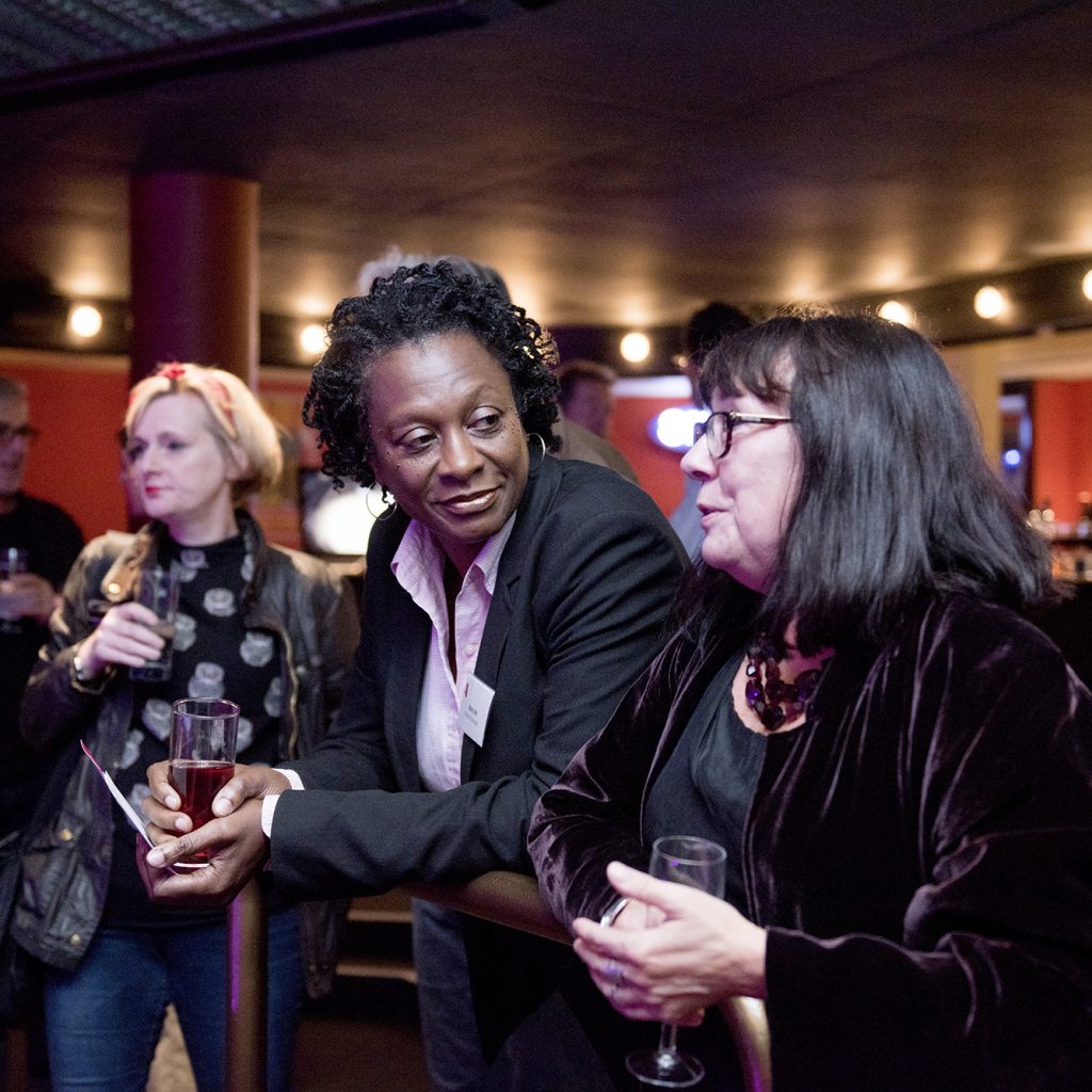 Two women lean on a balcony in the Albany theatre bar, holding drinks.