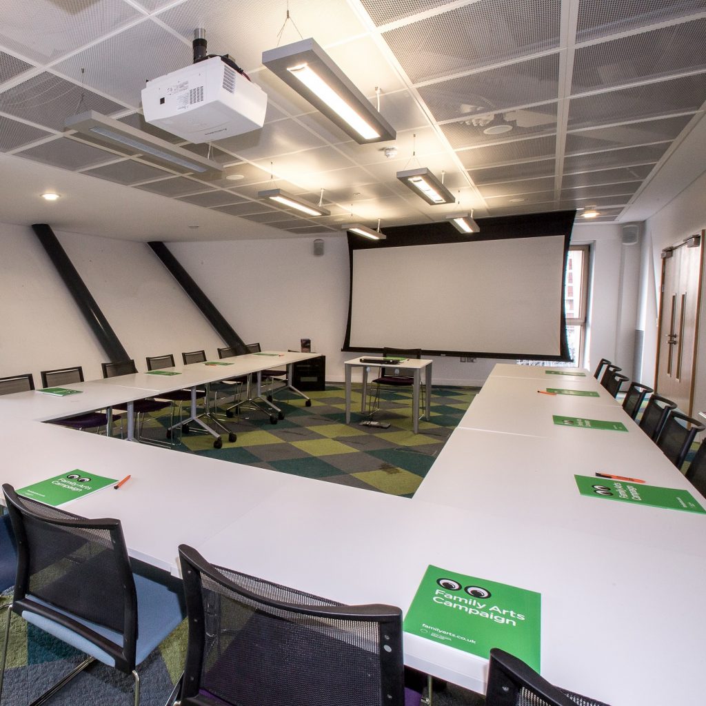 Large meeting room with big window on left wall and view of trees and Canada Water. Projector creen at far end.