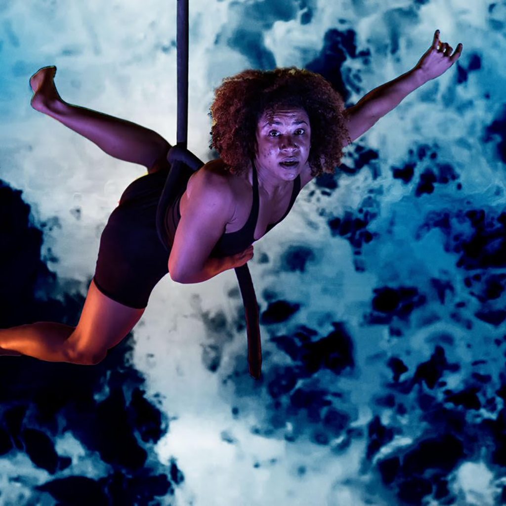 A female circus performer hangs from a rope which is around her waist. She is looking towards the camera.