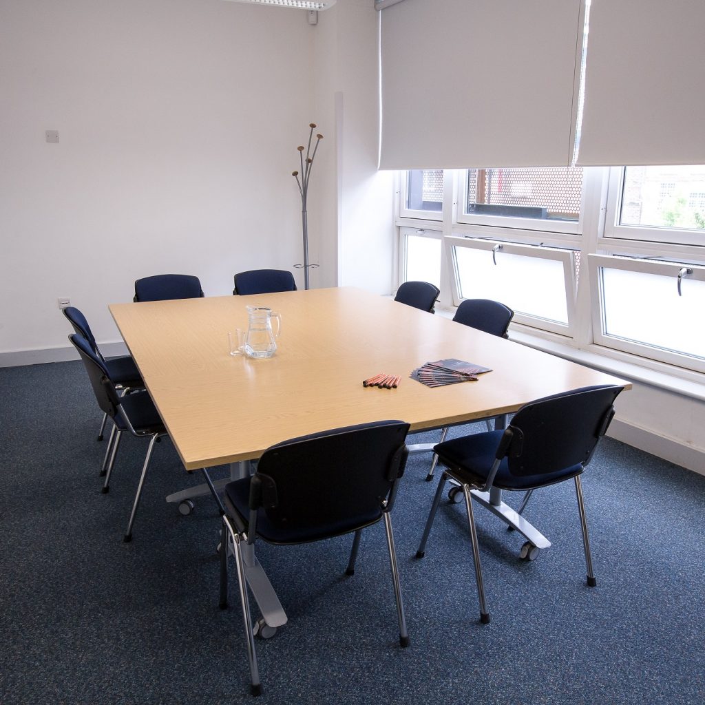 A small, bright meeting room with a large windo on the right and tables and chairs in boardroom style for 8