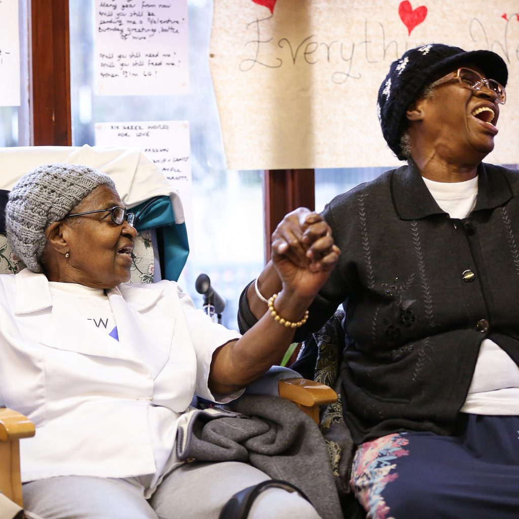Image shows two mature women of colour seated in comfy chairs holding hands and singing. There's a window with handwritten poetry and drawings pinned to it
