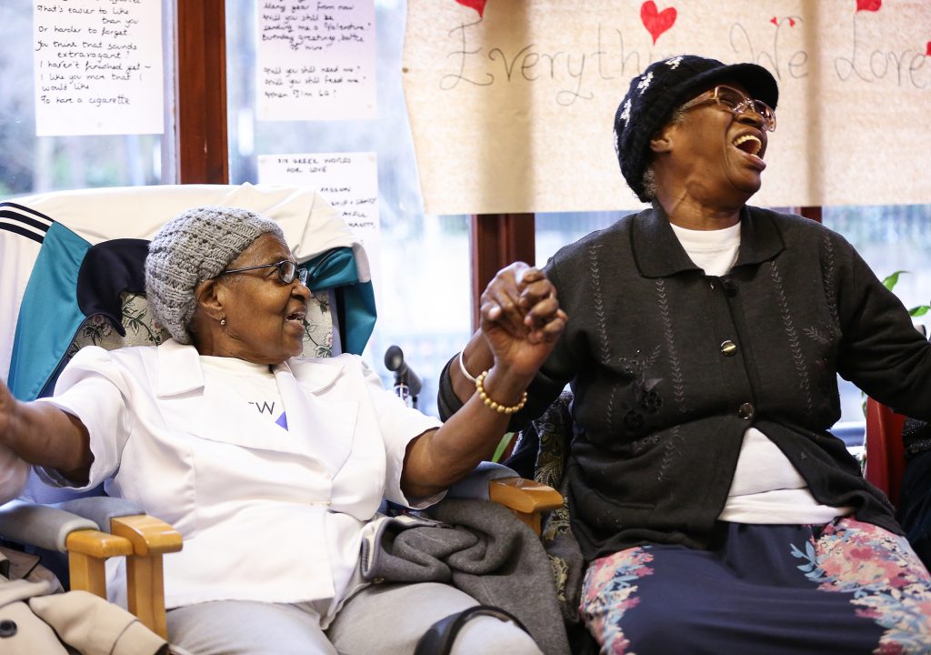 Image shows two mature women of colour seated in comfy chairs holding hands and singing. There's a window with handwritten poetry and drawings pinned to it