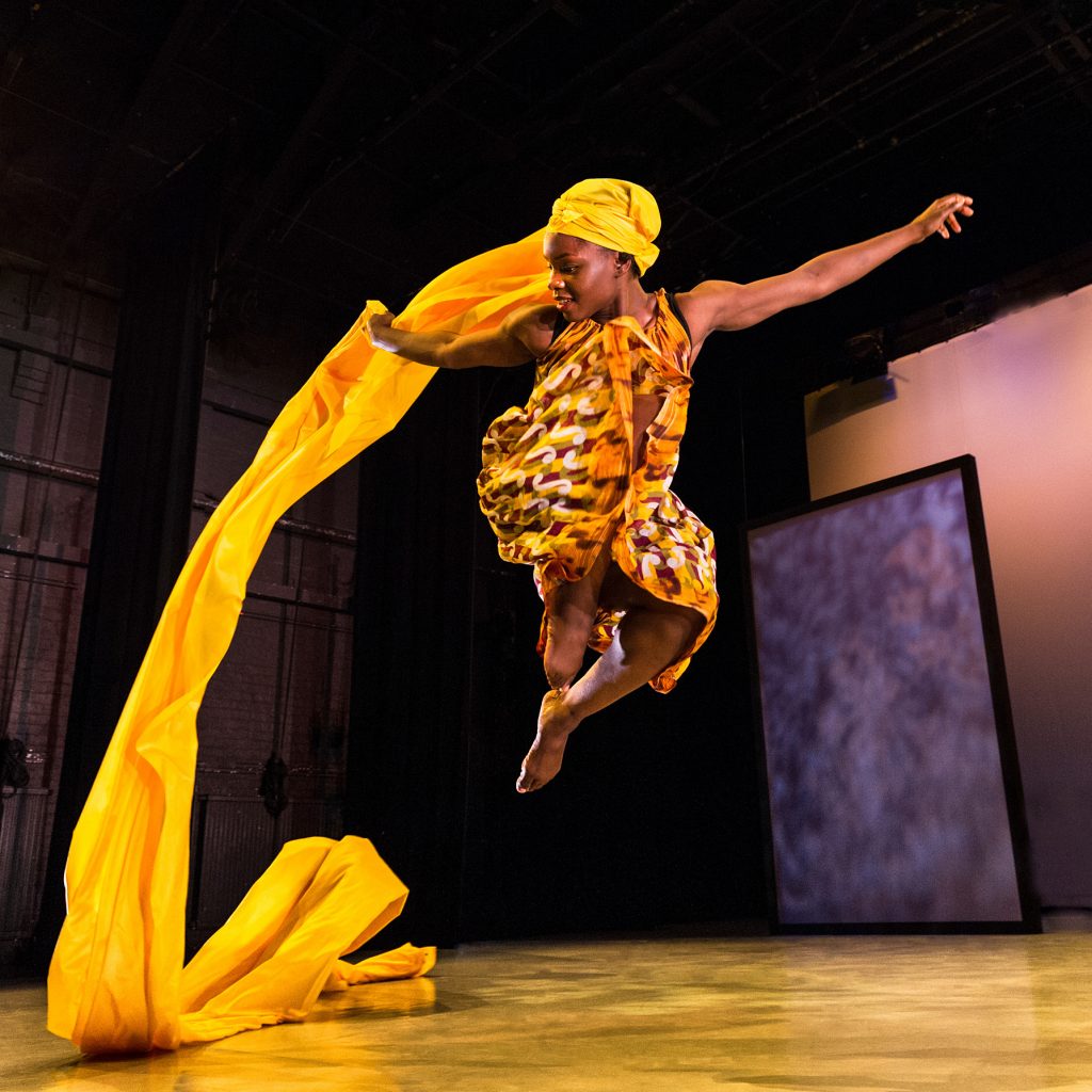 A female dancer with a bright yellow head wrap jumps in the air. The fabric flows out behind her from the wrap.
