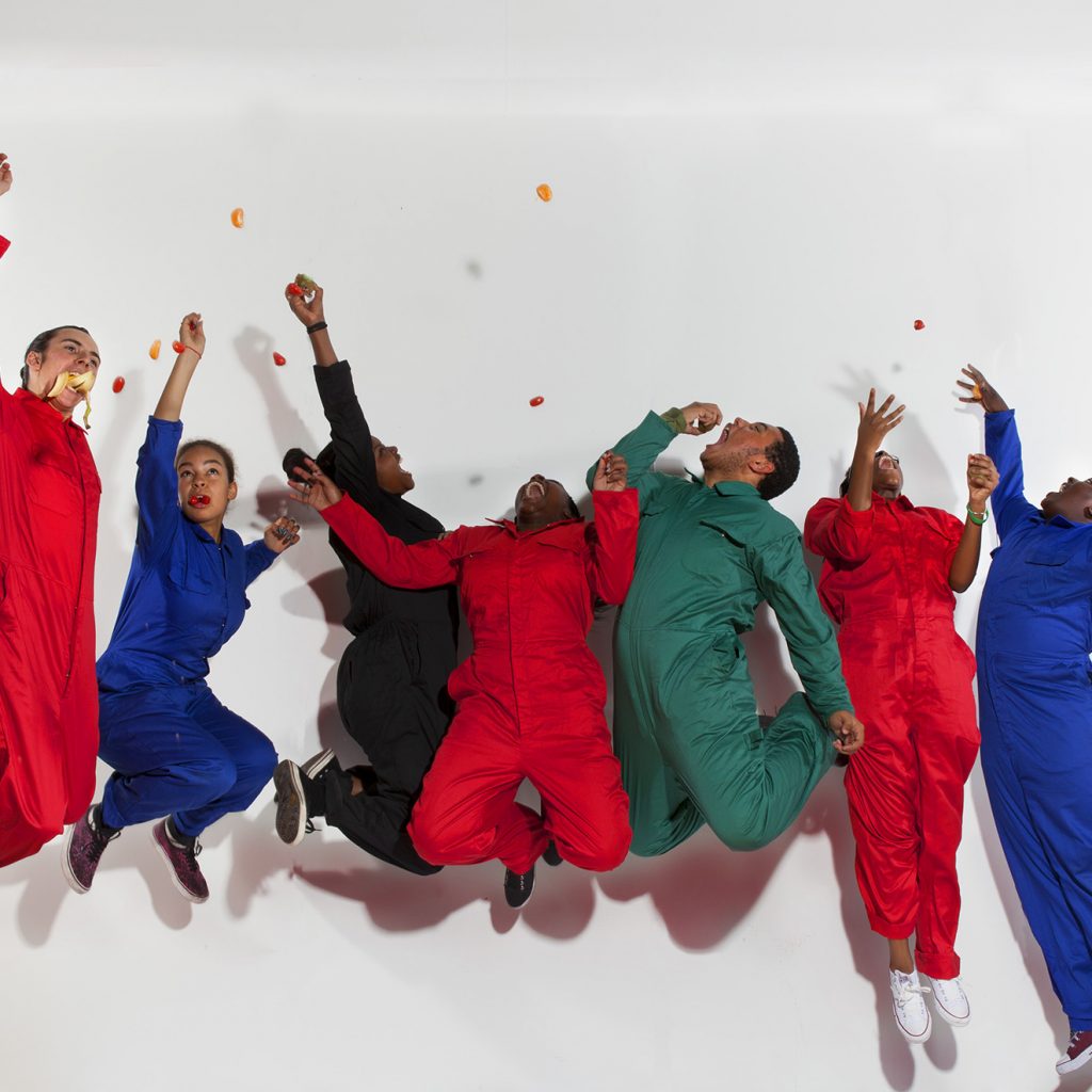 Seven young people in bright boiler suits leap in the air against a white background.