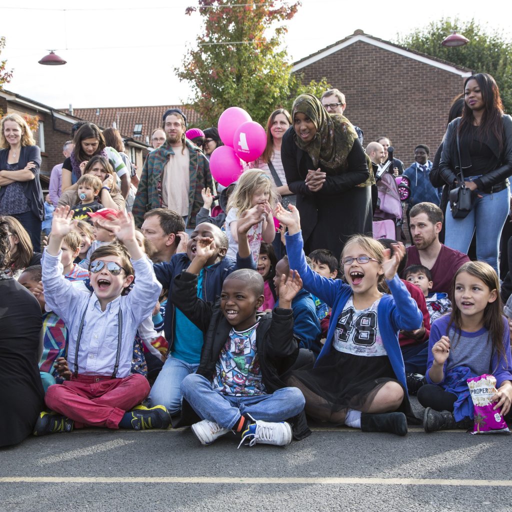 Large group of people of all ages, smiling and clapping as they watch a performance on Douglas Way, Deptford