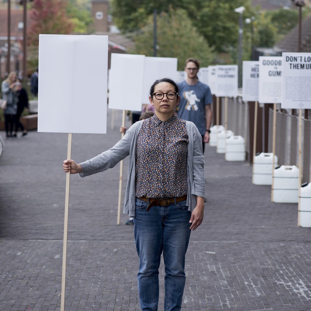 A woman stands on a cobbled street holding a blank plackard. Others stand in-line behind her.