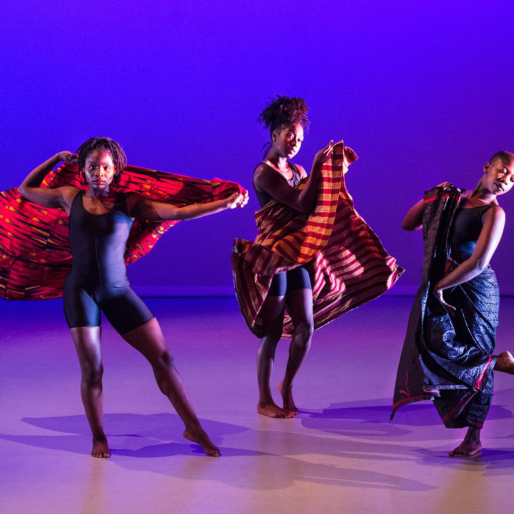 Three black, female dancers with head wraps under blue and purple lighting.