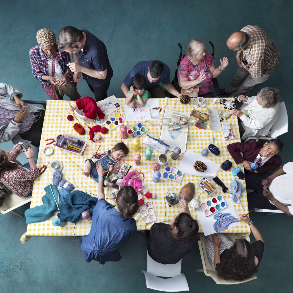 View of a table from above covered in craft materials with a baby lying on it and people of all ages stood and sat around it.