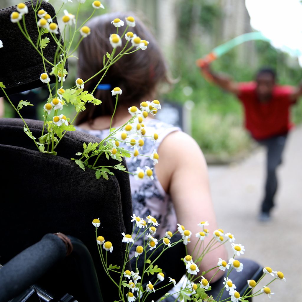 A woman in a wheel chair with flowers in it, wheels towards a man with a skipping rope on a street.