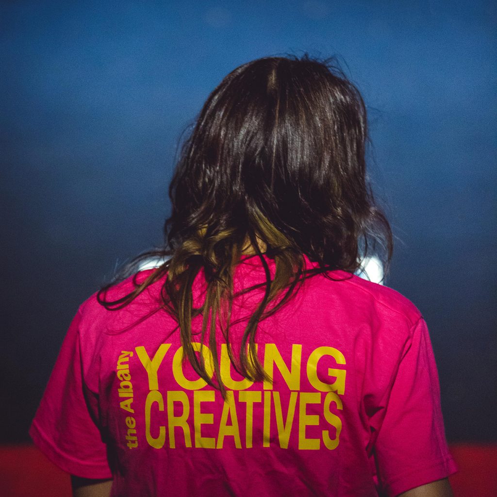 Person with shoulder length brown hair, seated with back to camera wearing a bright pink t-shirt with Young Creatives written on it in bright yellow.