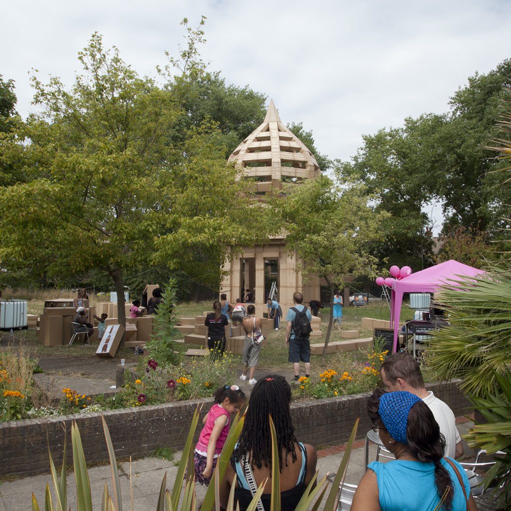 People on the patio in the Albany garden with a huge cardboard tower being built by members of the public in the background for a project called The People's Tower