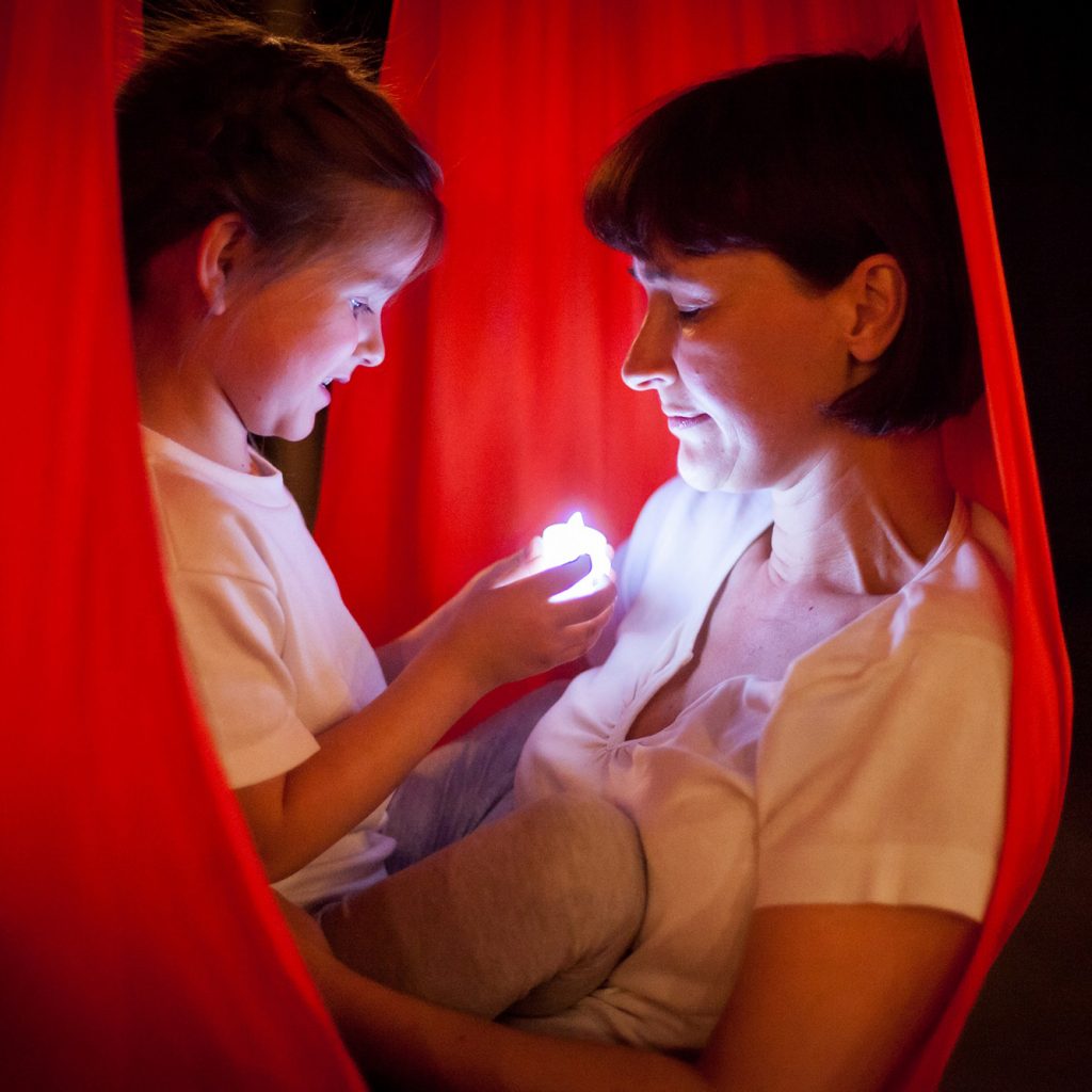 Mother and daughter seated enclosed in red fabric pod with glowing light.