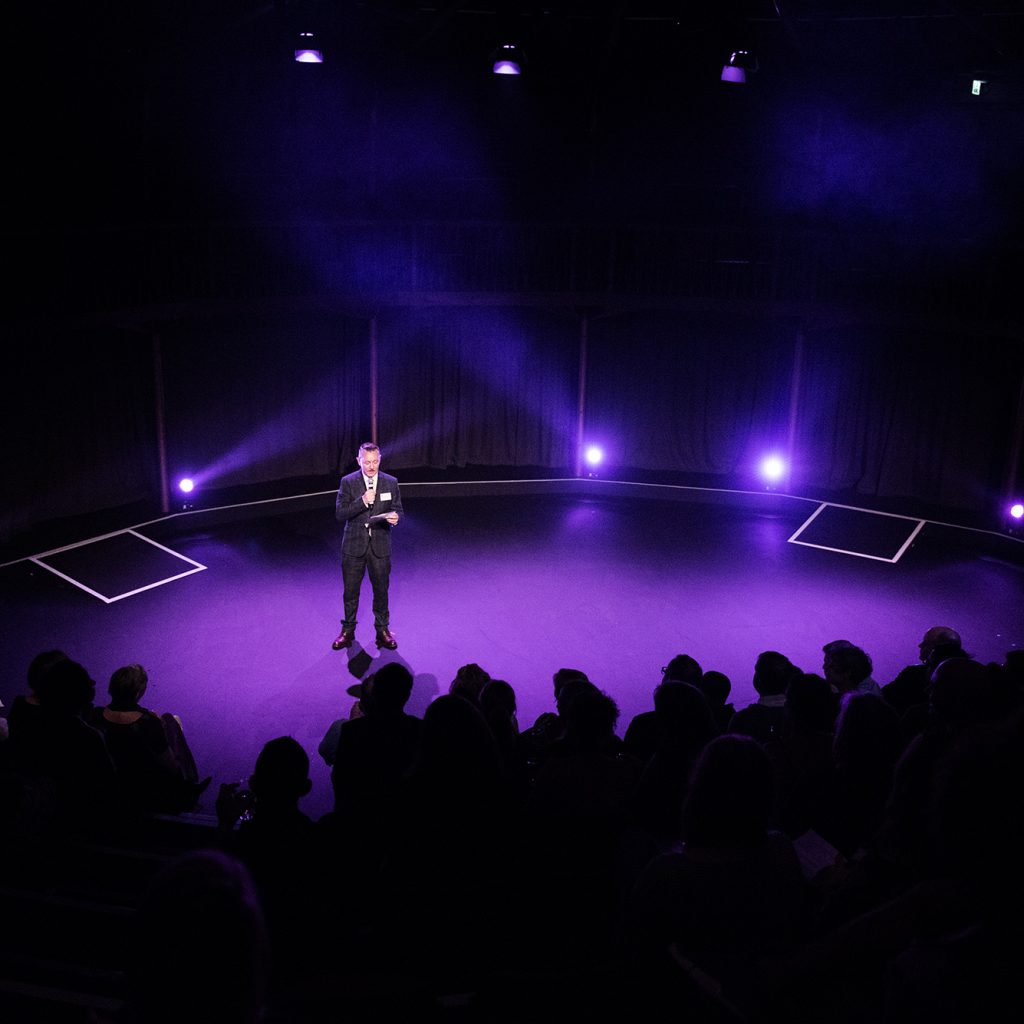 Image from the back of the Albany theatre. Man in suit stands on stage with microphone, full audience silhouetted in foreground.