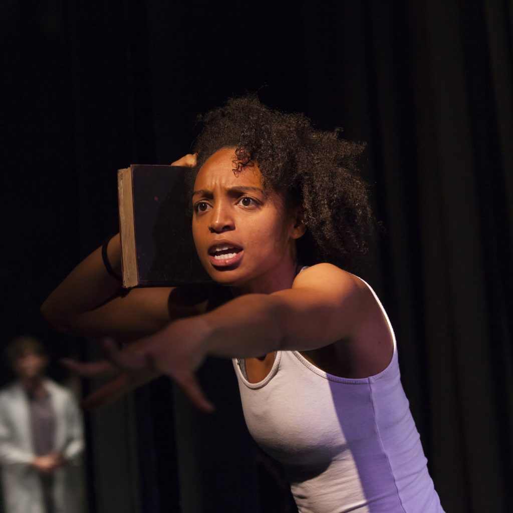 A young black woman wearing a white vest reaches towards the camera. She looks upset or angry but powerful.