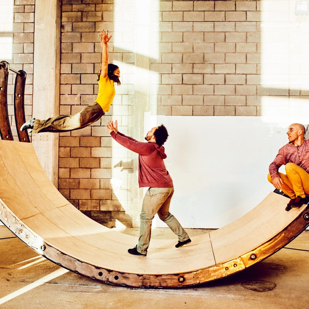 A huge, curved wooden seesaw with a woman leaping from one end of it into a man's arms.