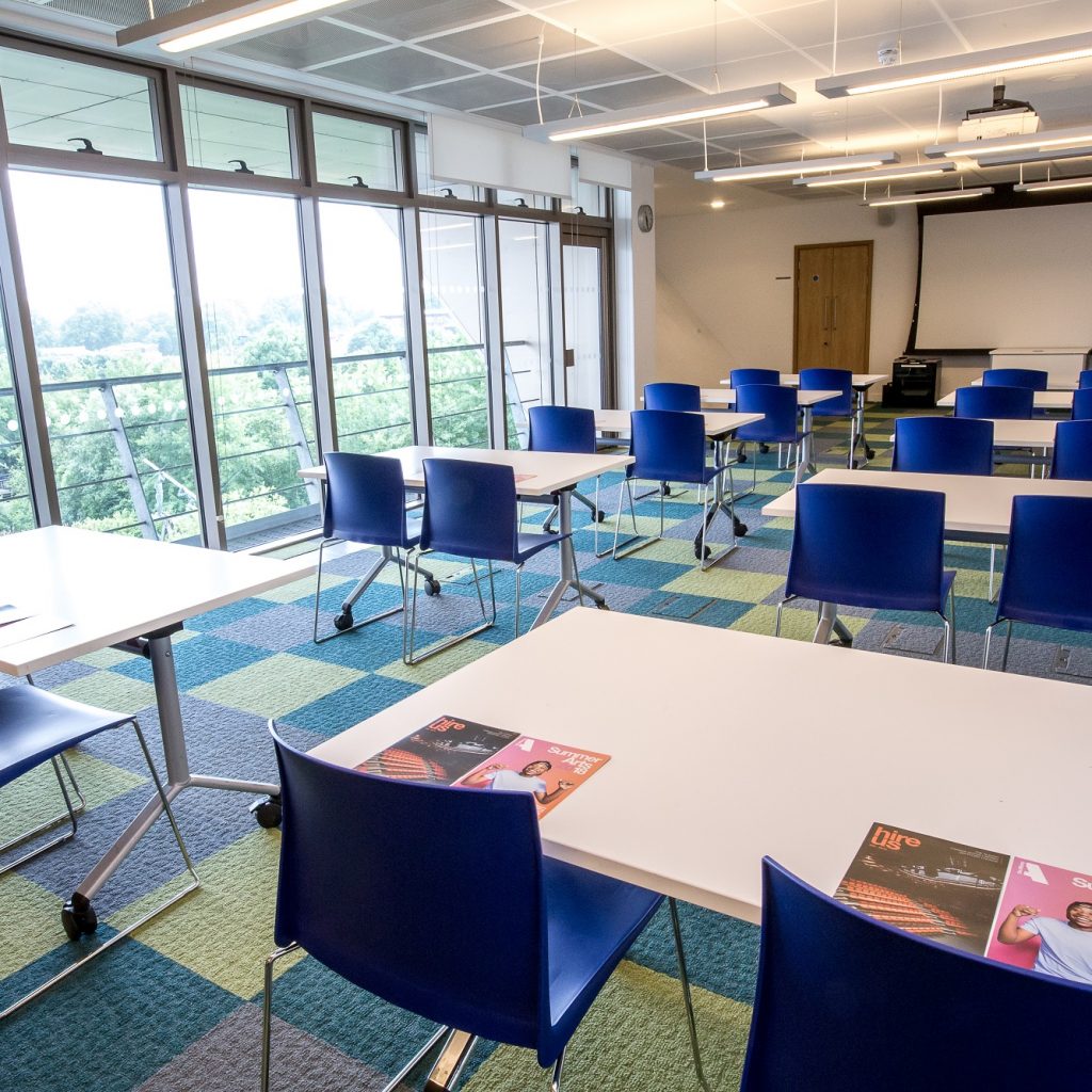 A bright meeting room with tables in set out like desks in a classroom and a white projector screen at the far end.