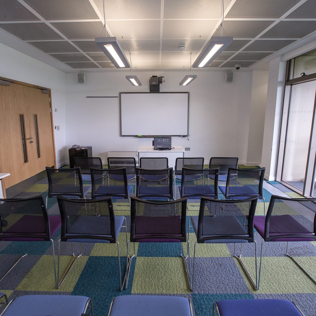 Large, bright meeting room with big window and chairs in rows and projector screen at far end