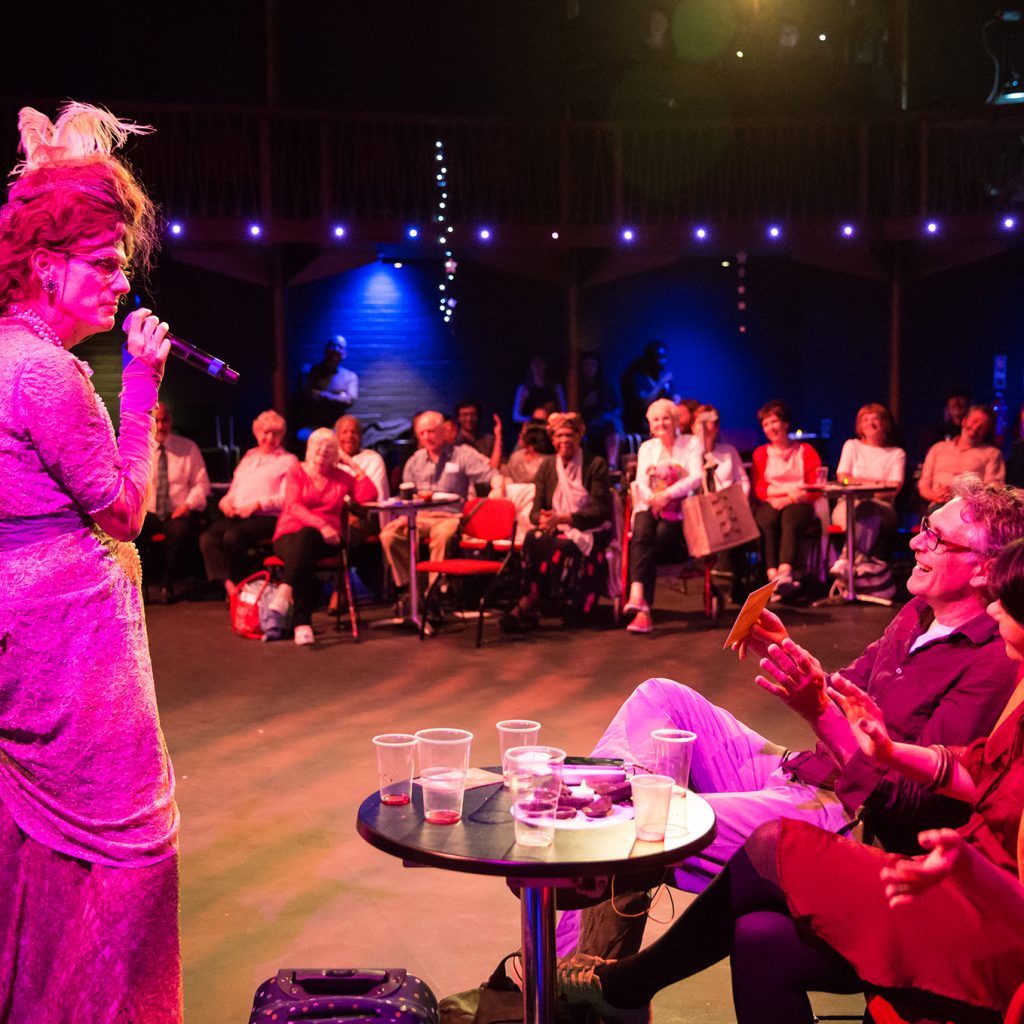 Chris Green dressed as mature woman with large hair and a full length lace dress (character is Ida Barr) stands with a microphone, speaking to an audience at cabaret tables.