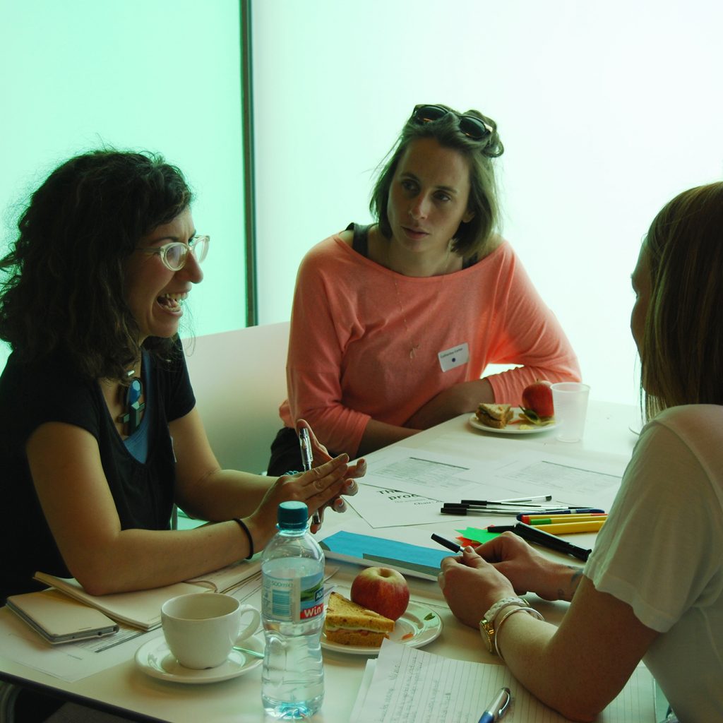 Four adults in casual work clothes sit around a table with drinks and snacks, talking.