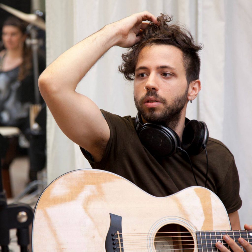 A man in a black t-shirt holding an acoustic guitar is in thought