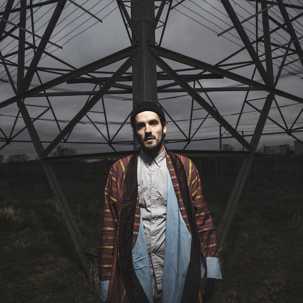 Dizraeli stands in front of a huge electricity pylon under a moody, grey sky.