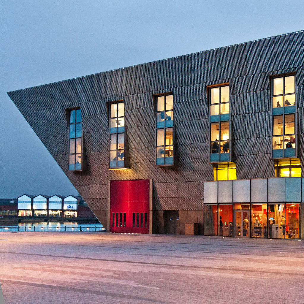Large, modern, asymmetric building with steel cladding and warmly lit windows.