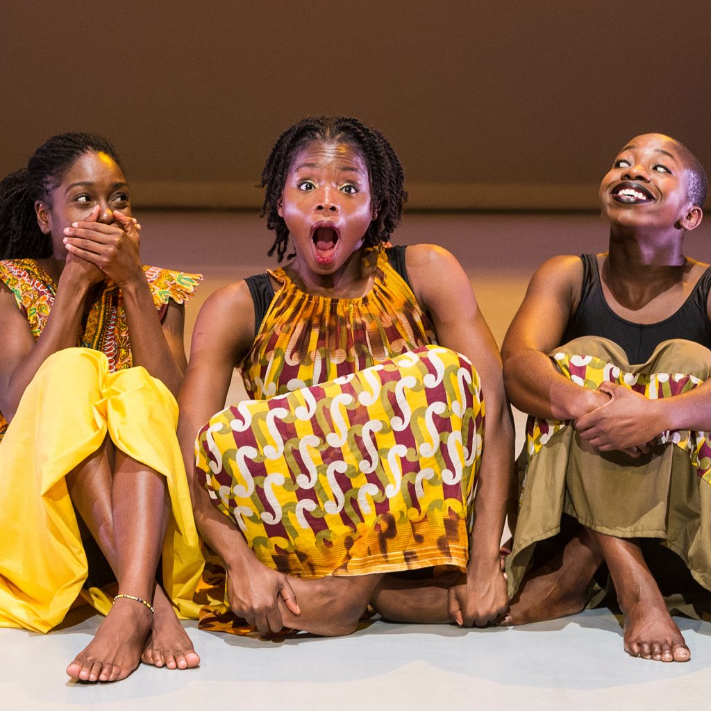 Three young women of colour in yellow and black dresses sit side by side on the floor. They are laughing or smiling.