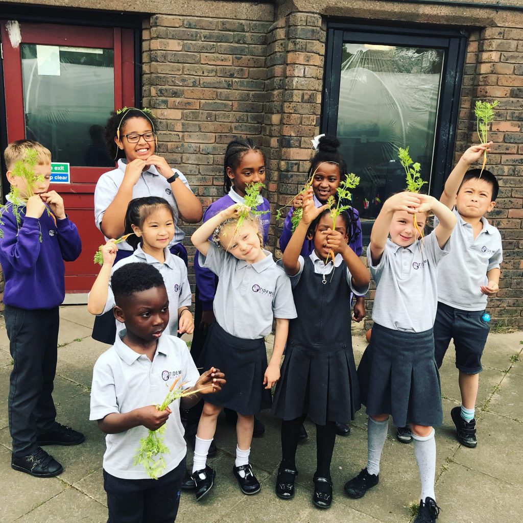 A group of 5 or 6 year olds pose in front of a wall, holding up carrots. They are wearing school uniform and look happy and proud.