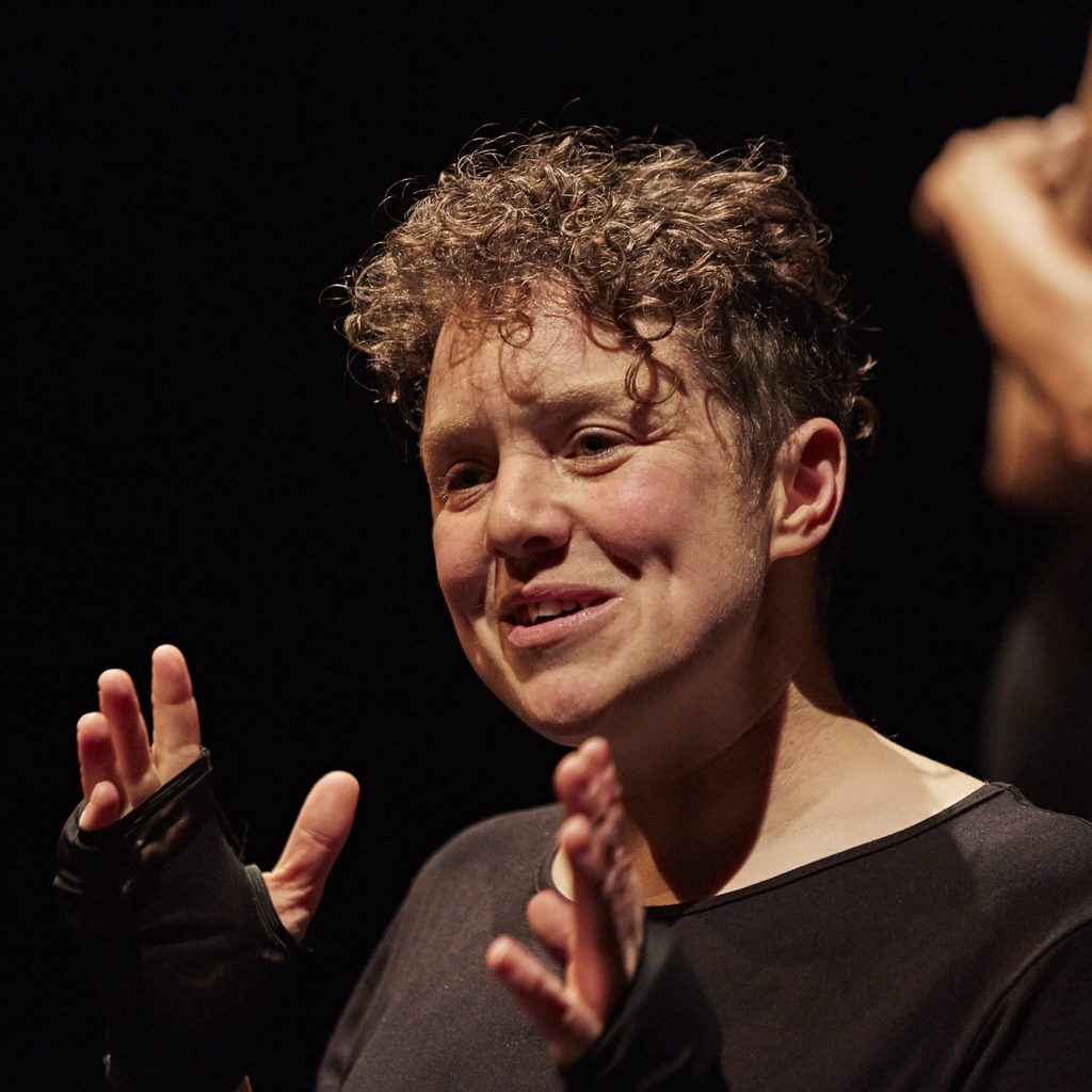 Picture of Jess Thom - a white woman in her 30s with short curly hair, wearing a black T-short and holding her hands up as she talks animatedly.