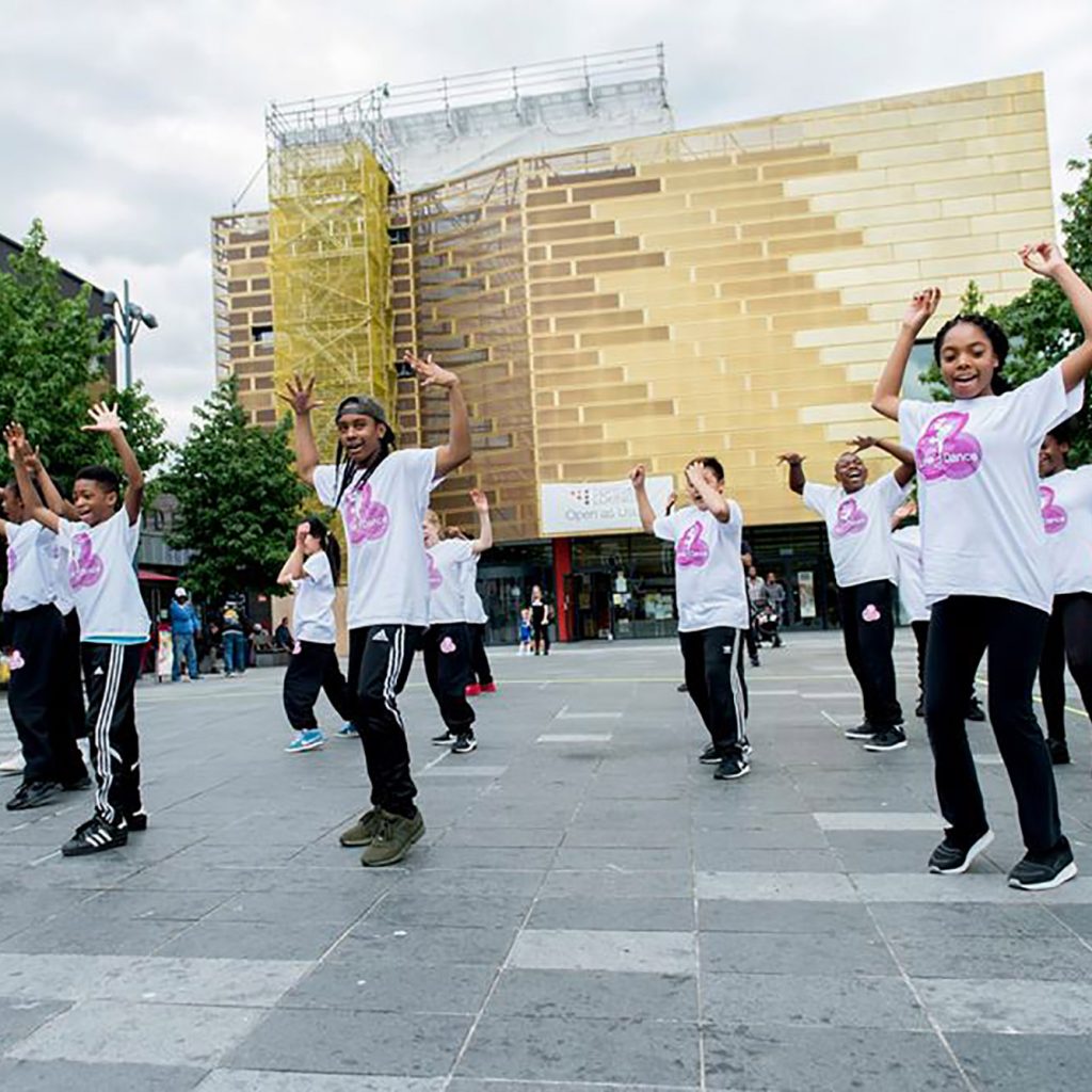 Large concrete open space in front of Deptford Lounge with trees, benches and young dancers performing