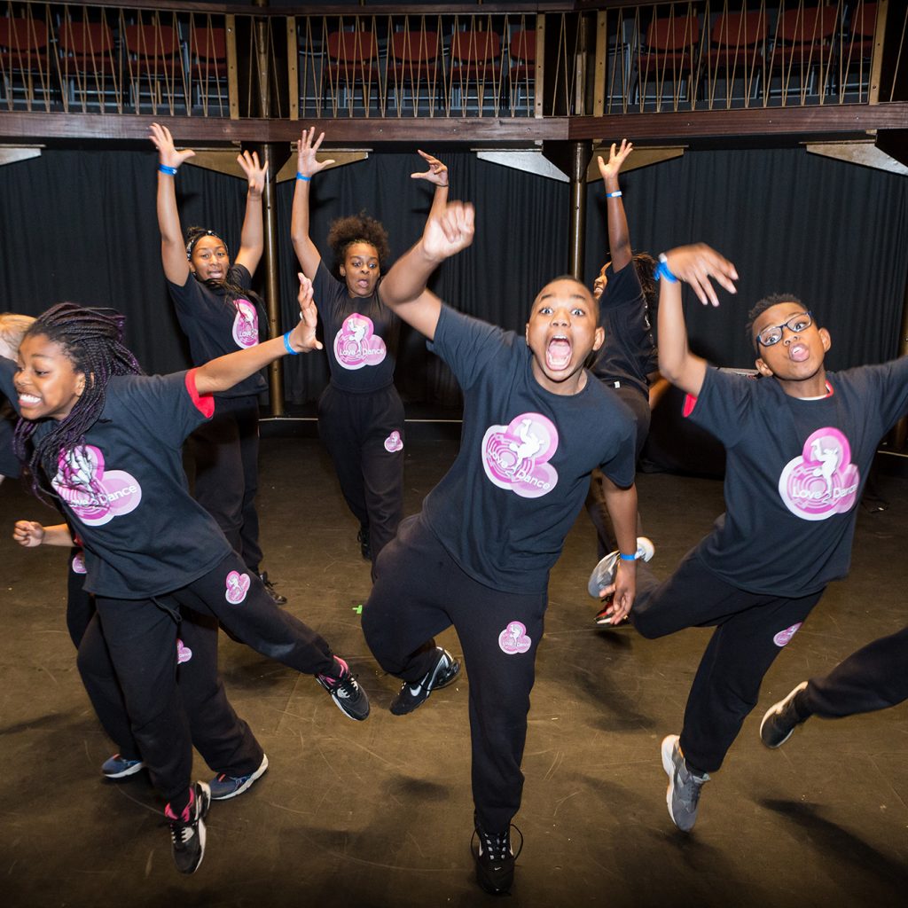 Young performers in black and pink T-shirts dance on the Albany stage.