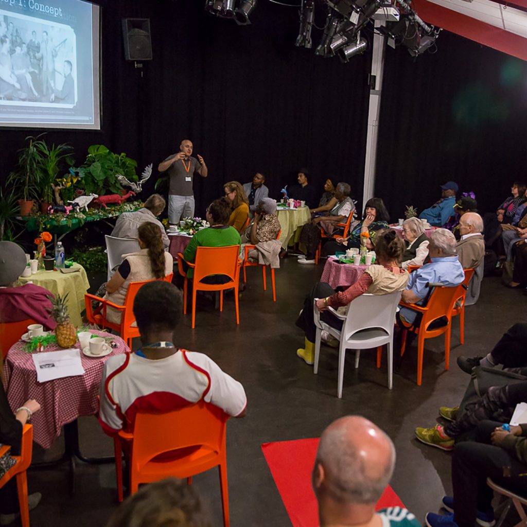 Lots of people sit in a large performance studio around tables and in a row of seats. They are about to watch a film on a large screen at the front of the room. There is a display with sculptures and a model forest at the front of the room.