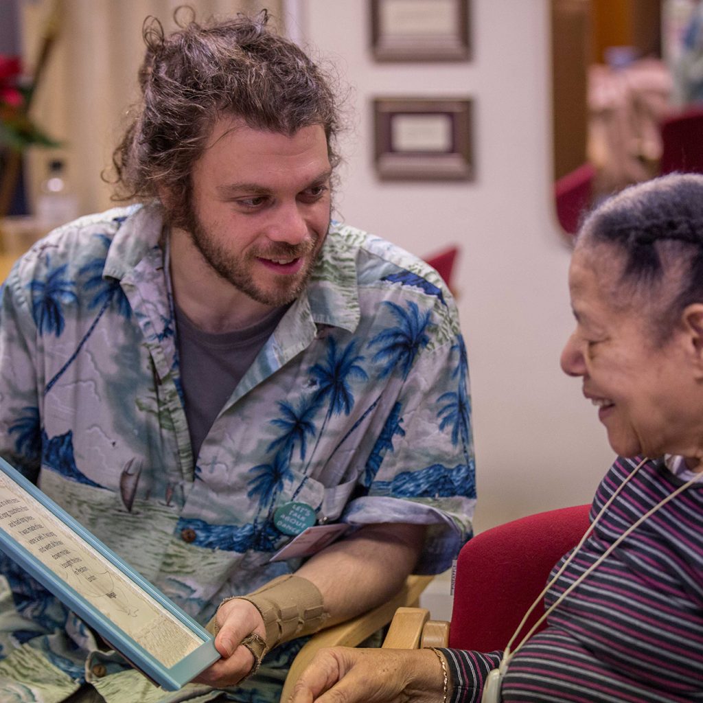 A younger man shows an older lady something in a blue photo frame. They both look happy. They are both sitting down.
