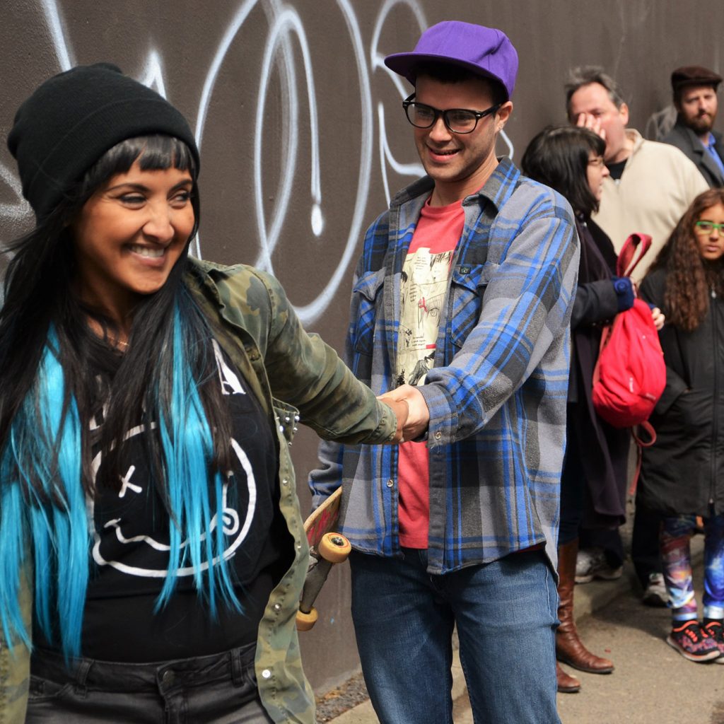 A young woman in a black woolly hat with black and bright blue hair grins as she leads a young man by the hand up an urban street. A small crowd of all ages look on.