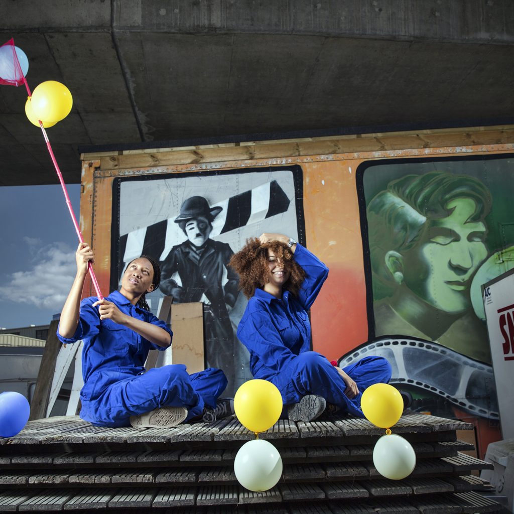 Two young people in blue boiler suits sit atop a pile of wooden pallets. There is a snack truck beside them and there are yellow and white balloons.