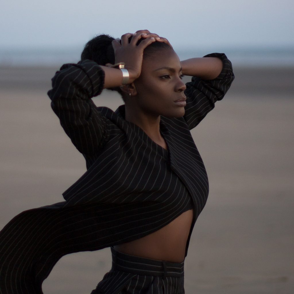 A beautiful black woman in a pinstripe suit with the tails blowing behind her, looks into the distance on a beach.