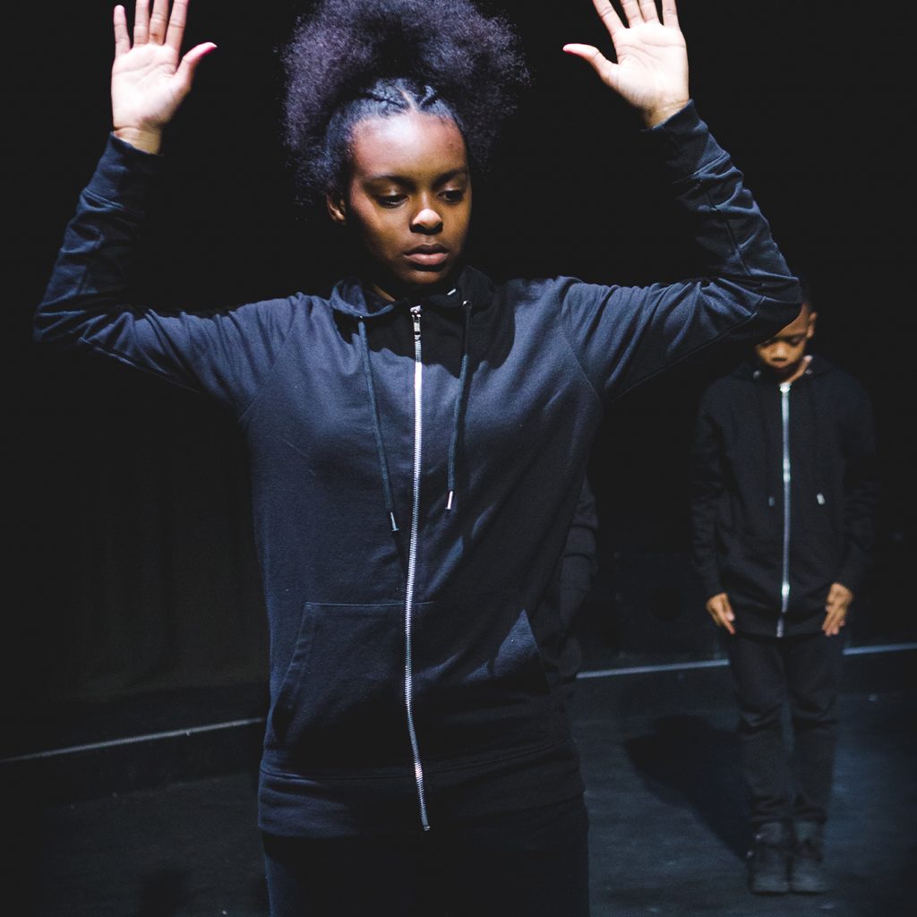 Young dancer in a black tracksuit with her arms in the air, performing in The Cloud on the Albany stage.