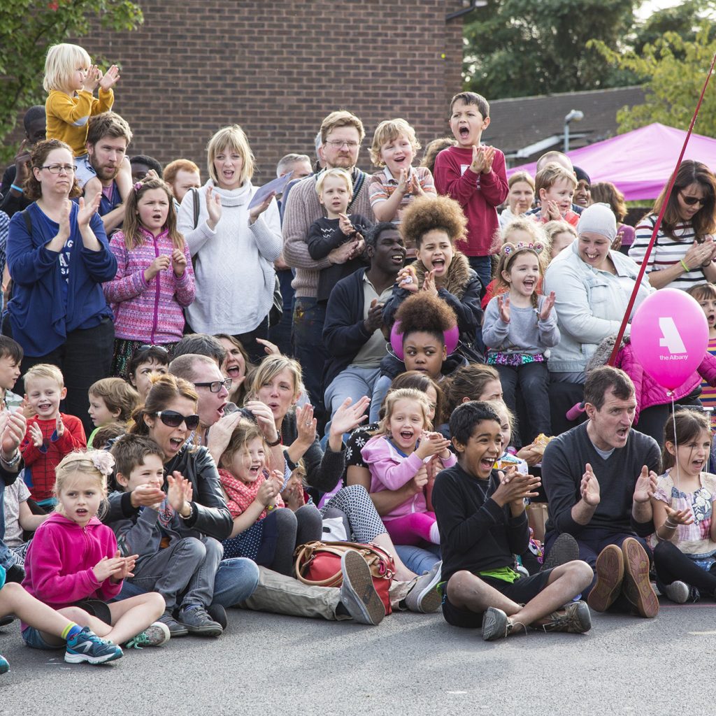 Large group of people of all ages, smiling and clapping as they watch a performance on Douglas Way, Deptford