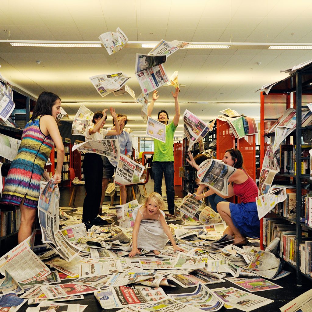 Children and adults throw newspapers in the air in a modern library.
