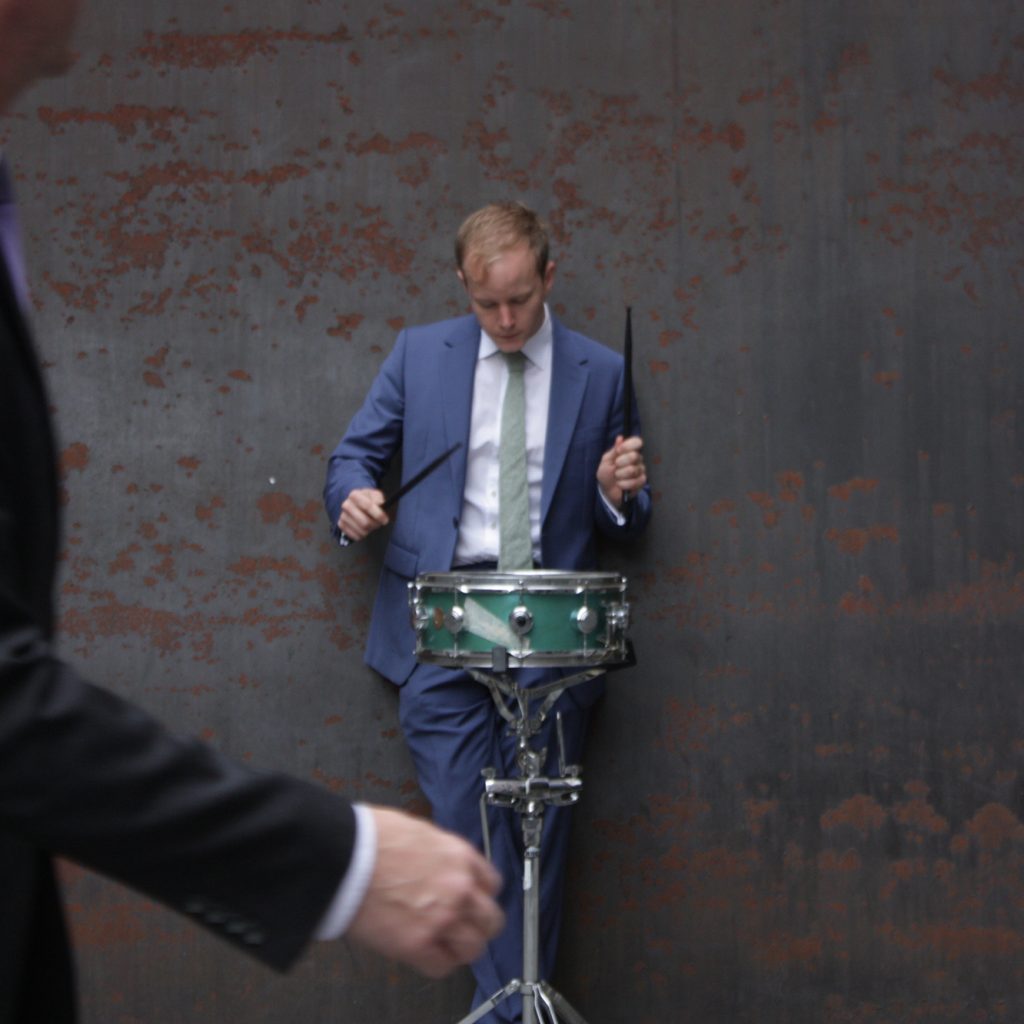 Suitman Jungle: man in a suit, standing up, playing a small drum.