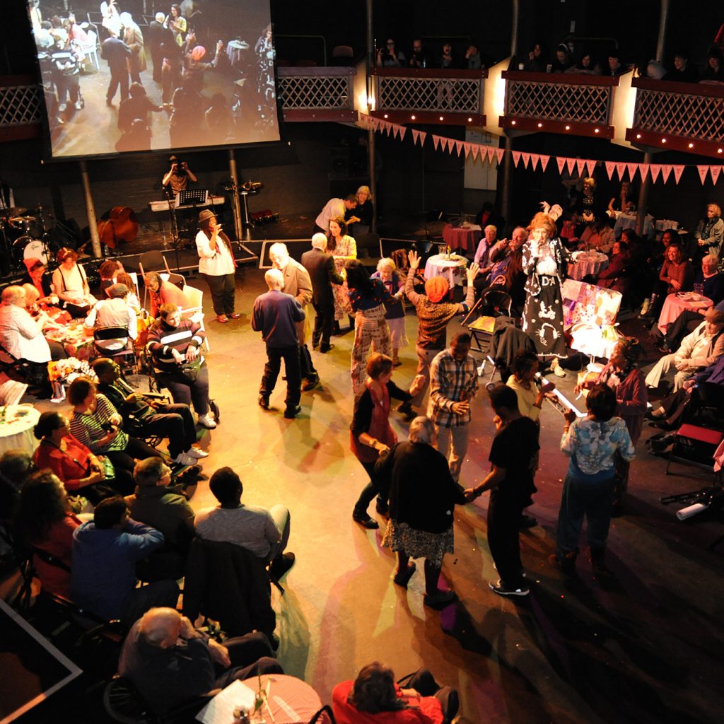 A theatre filled with people of all ages dancing, sitting at tables, with a big screen at the back.