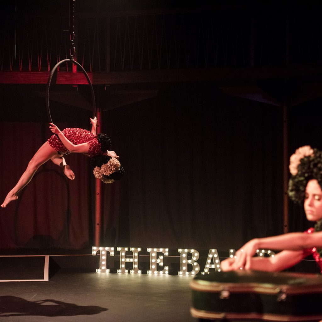 Woman in foreground, seated behind guitar case, woman in background hanging off aerial circus hoop. Both in red sequin dresses with extravagant black wigs.