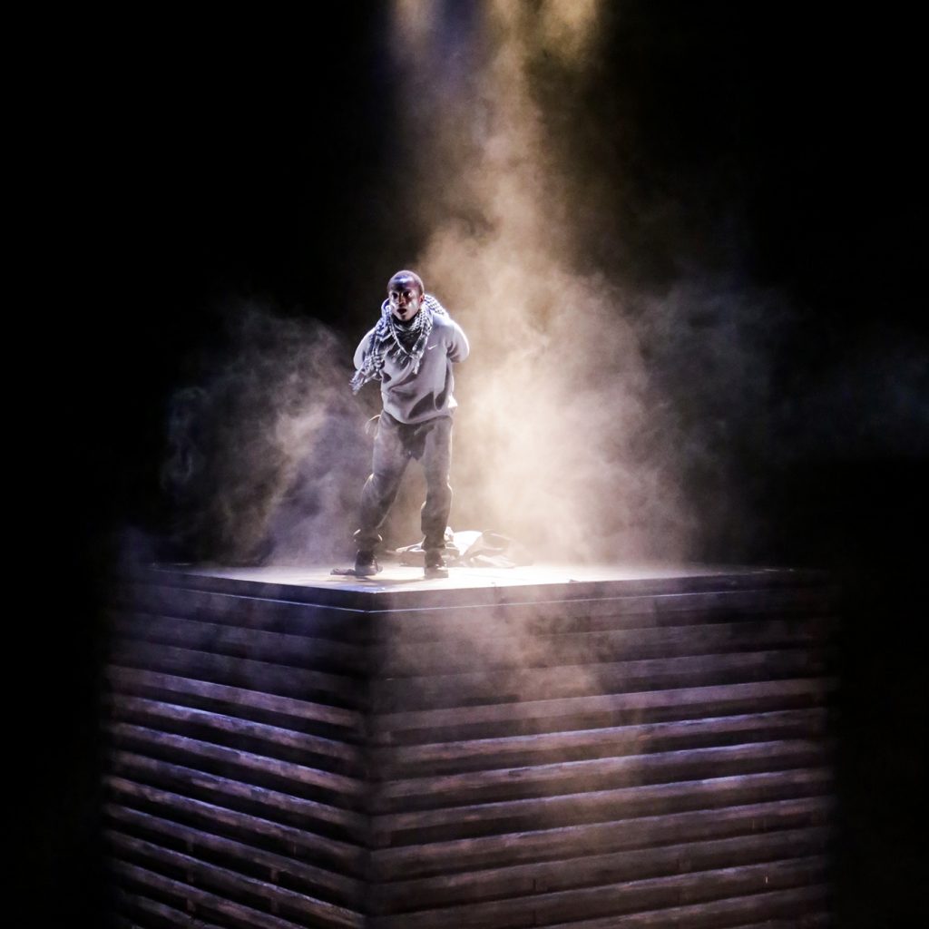 A male actor stands wreathed in smoke on top of a huge wooden box. He is lit from above.