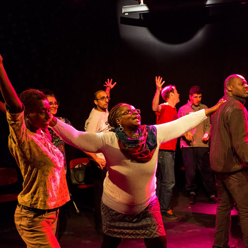 A group of people in a room, in a loose circle. Most have with their arms raised, they look like they're dancing.