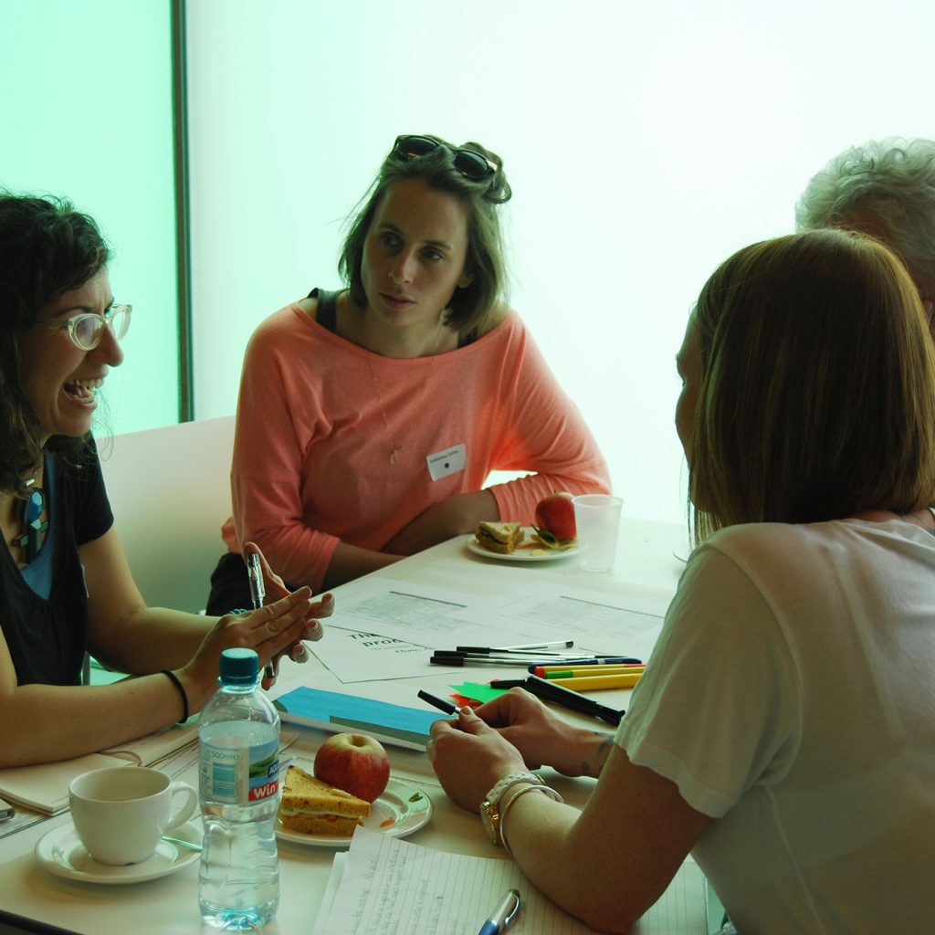 Four adults in casual work clothes sit around a table with drinks and snacks, talking.
