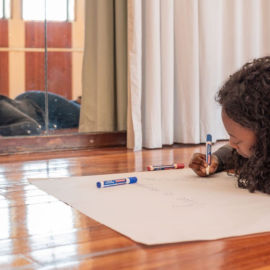A young black woman lies on the floor of a dance studio, writing on a big piece of paper. You can see her reflection in a mirror. She is wearing a great top and jeans.