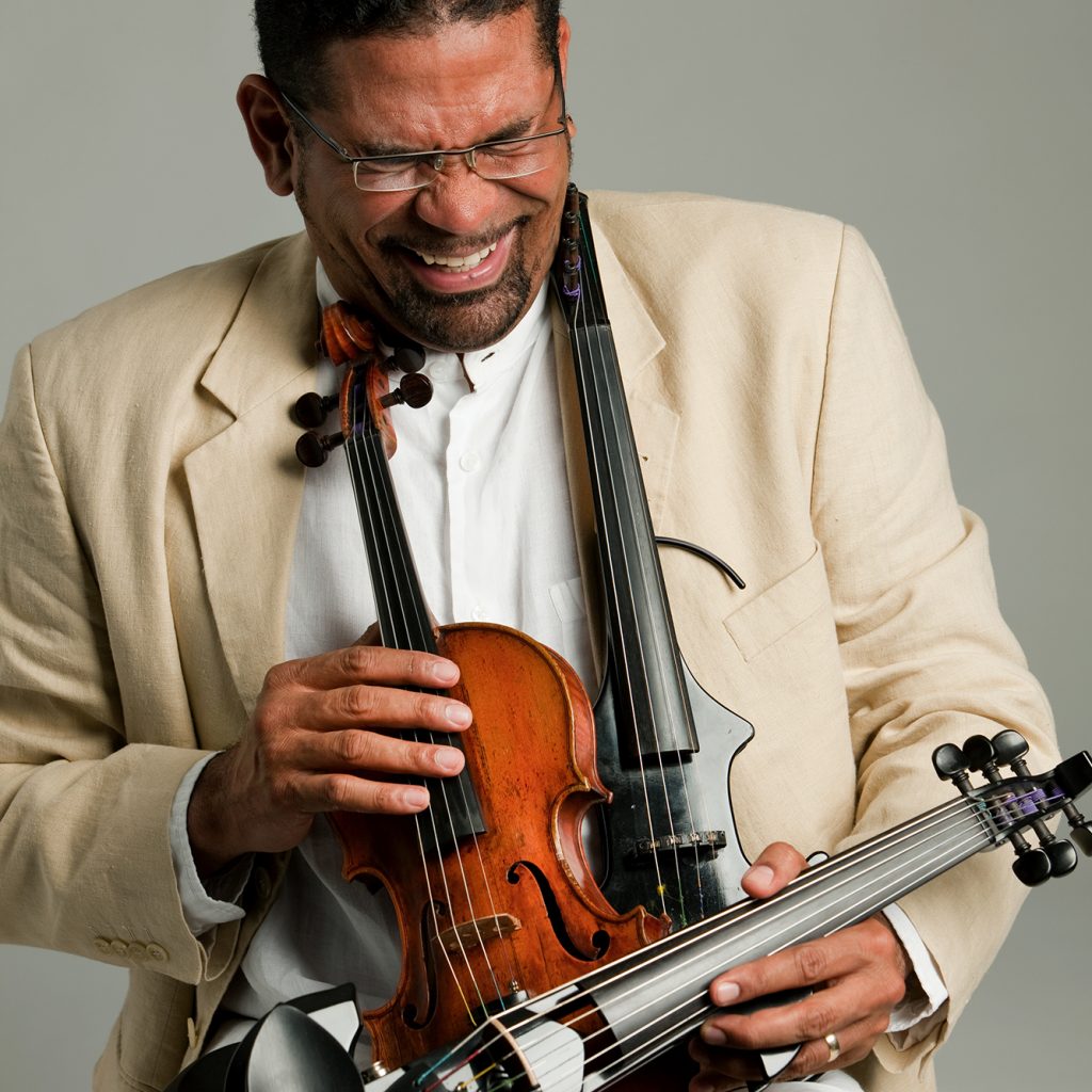A Latino man in a cream suit, white shirt and glasses laughs with his face scrunched up. He is holding several violins.