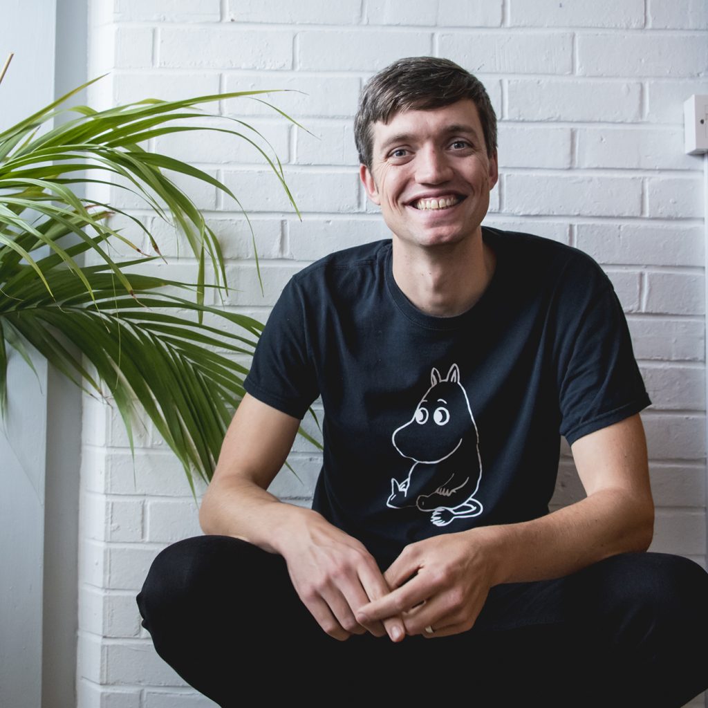 A young man wearing black crouches down and smiles at the camera in front of a white wall.