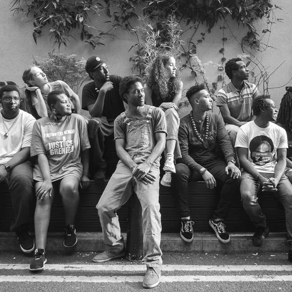 Black and white photo of a large group of adults in their 20s siting outside on a bench with a wall with plans growing up it behind them. They are all looking to the right.