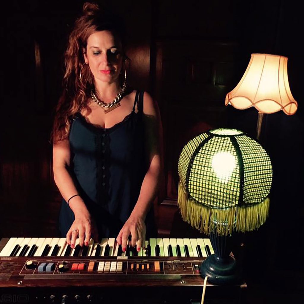 Image of a white woman with auburn hair and chunky jewelry in a blue dress. she is standing playing a keyboard with a lamp on her right and anther lamp behind her.